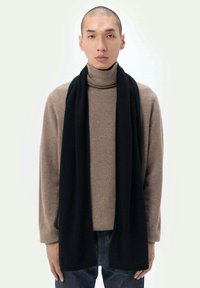 Svart halsduk som ligger draperad över en brun polotröja, med en mjuk textur och en enkel, avlång design. En enkel bakgrund.