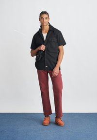 Camisa preta de manga curta com dois bolsos no peito, calças bordeaux e sapatos laranja com atacadores, em pé sobre um tapete azul contra um fundo branco.