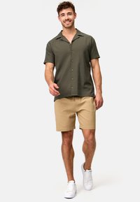 Camisa de oliva de manga corta con botones, combinada con short beige, llevada con zapatillas blancas. Textura suave y corte casual.