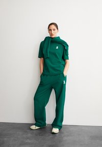 Grünes Kurzarm-Oberteil mit weißem Logo, kombiniert mit passenden, lockeren Hosen. Ein Pullover ist lässig um die Schultern gelegt. Leichte Schuhe runden das Outfit ab.