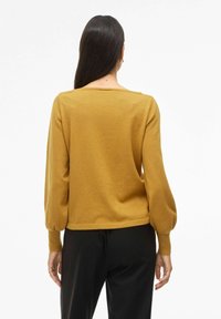 Maglione di colore senape in maglia con scollatura ampia e maniche lunghe a palloncino; texture liscia, orlo all'altezza della vita. Visibili pantaloni neri.