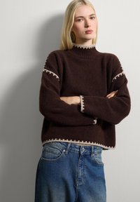 Femme blonde avec les bras croisés, portant un pull marron foncé avec des bordures claires et un jean bleu, se tenant contre un mur gris clair.