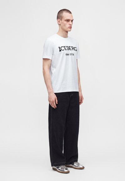 Νέος άνδρας με λευκό t-shirt Iceberg, μαύρο παντελόνι με φαρδιά πόδια και ασημένια αθλητικά παπούτσια, στέκεται μπροστά σε απλό ανοιχτό φόντο.