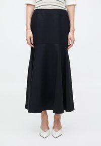 Woman wearing a black high-waisted, flared midi skirt with a white top and white pointed-toe heels, standing against a plain background.