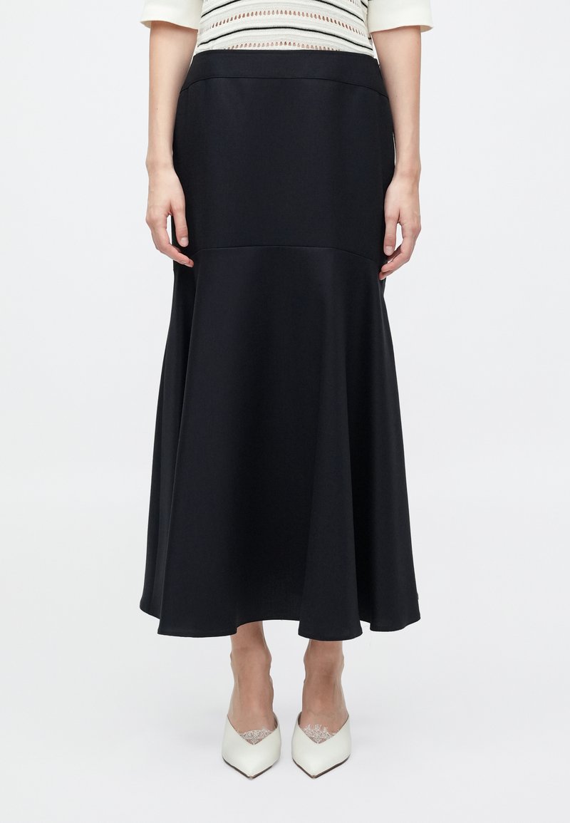 Woman wearing a black high-waisted, flared midi skirt with a white top and white pointed-toe heels, standing against a plain background.