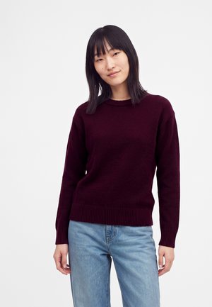Jonge vrouw met stijl zwart haar, gekleed in een donkerpaarse trui en lichtblauwe spijkerbroek, staand tegen een effen witte achtergrond.