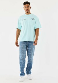 Jeune homme portant un t-shirt oversized bleu clair et un jean bleu avec des motifs en texte blanc, se tenant contre un fond blanc uni.