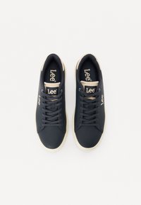 Baskets noires en matériau synthétique avec une texture lisse. Dotées de lacets crème et d'un logo beige sur la languette, design minimaliste.
