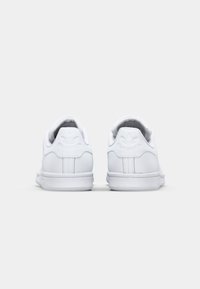 Zapatillas de cuero blanco con textura suave, punta redondeada y cuello acolchado. Incluye logotipo en relieve en el talón y suela de goma plana. Vista trasera.