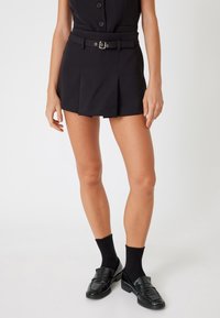 Pantalones cortos negros ajustados con un cinturón, que presentan pliegues y una textura suave. Combinados con calcetas negras y mocasines brillantes.