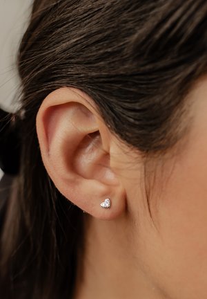 Silver heart-shaped earring with small clear gemstones, placed in the lobe of a dark-haired ear, against a neutral background.