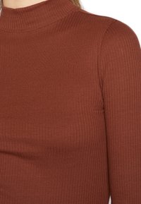 Rippstrick-Turtleneck-Oberteil in Rostfarbe aus dehnbarem Stoff. Figurbetontes Design mit langen Ärmeln und hohem Kragen.
