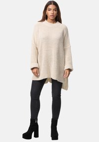 Beige gestrickter Oversize-Pullover mit einem gerippten Muster, weiten Ärmeln und einem High-Low-Saum, kombiniert mit schwarzen Slim-Fit-Jeans und schwarzen Plateaustiefeln.