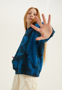 Felpa blue tie-dye in misto cotone, con tasca frontale, polsini e orlo a coste. Il modello posa con la mano alzata.