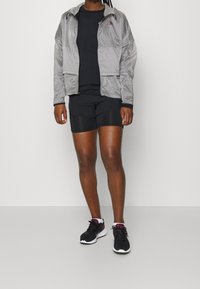 Lehká stříbrná větrovka s předním zipem, černé tričko a černé šortky, doplněné černými teniskami s fialovými akcenty.