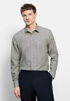 Uomo che indossa una camicia a maniche lunghe con bottoni con motivo astratto bianco e nero, abbinata a pantaloni blu scuro, in piedi con una mano in tasca.