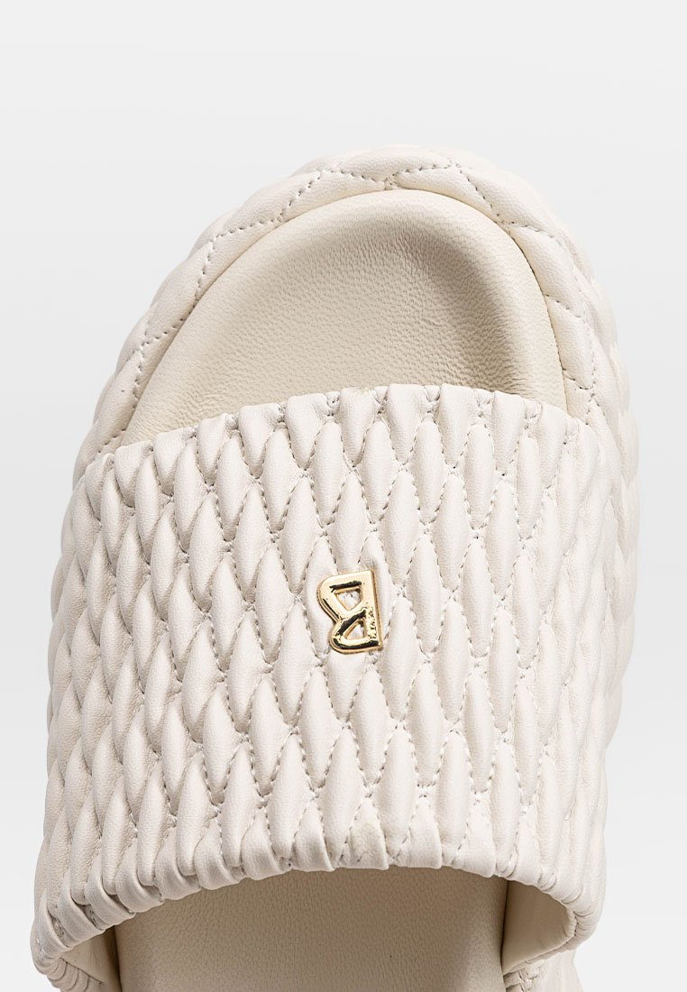 Beżowe, pikowane klapki z teksturowanym paskiem, z małym złotym emblematem "B" umieszczonym na górze na środku.