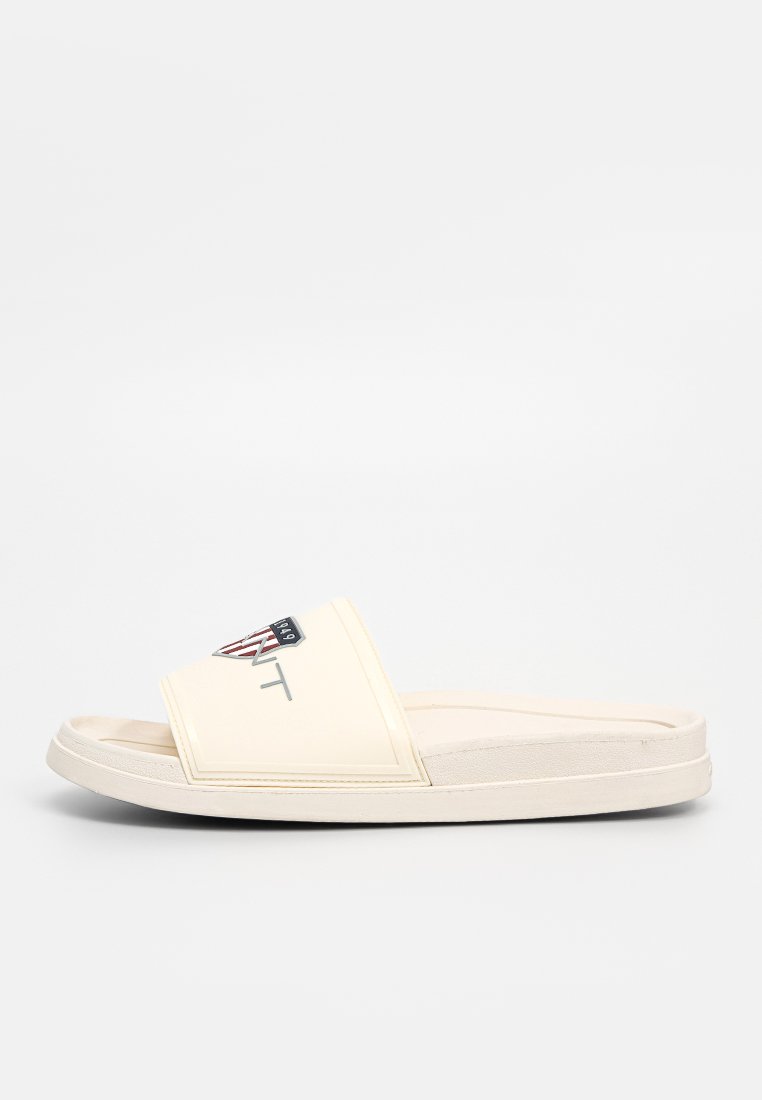 Riemensandale in Creme mit flachem Design. Verfügt über ein breites Oberband, strukturierte Akzente und ein Logo-Patch mit roten und schwarzen Details.