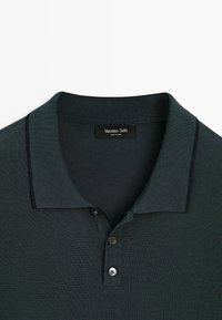 Mörkgrön polo t-shirt tillverkad av texturerat tyg. Har en ribbad krage, tvåknappsknäppning och kontrasterande piping längs kragen.
