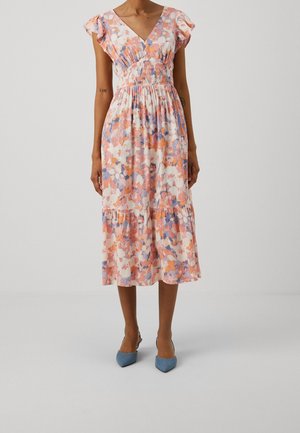 Vrouw draagt een bloemenmidi-jurk met wappermouwtjes, een geplooide taille en een gelaagde rok, gecombineerd met blauwe pumps met puntige neus.