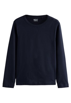 Žena s dlhými hnedými vlasmi, nosiaca malé kruhové náušnice, tmavomodrú blúzku s dlhým rukávom a modré džínsy, otočená chrbtom k jednoliatemu pozadiu.