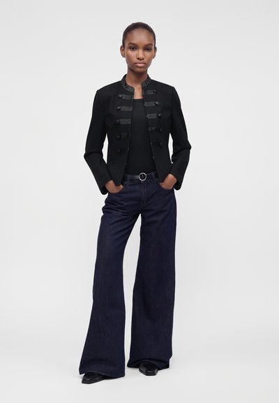 Femme debout, mains dans les poches, portant une veste noire de style militaire, un haut noir, un jean large bleu foncé et des chaussures noires.