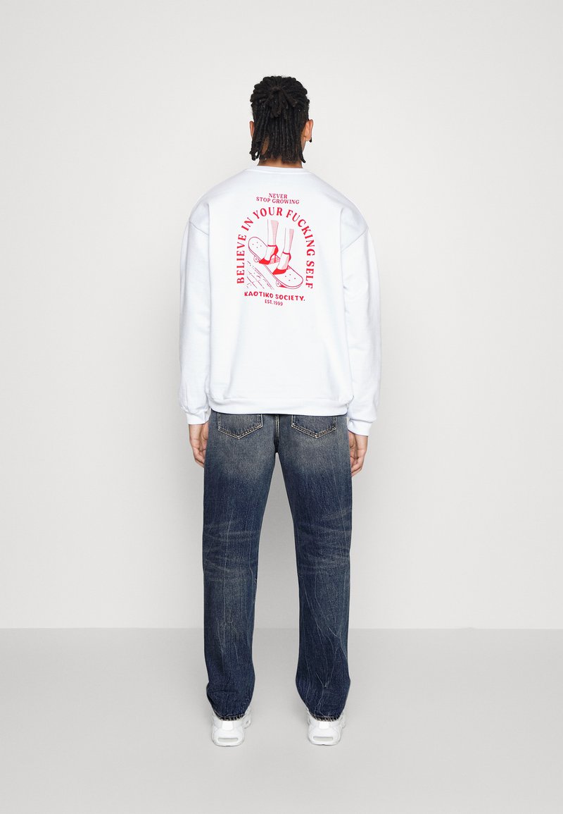 Weißes Sweatshirt mit rotem grafischem Text und Design auf dem Rücken, kombiniert mit dunkelblauen Jeans. Das Sweatshirt hat eine lässige Passform und gerippte Bündchen.