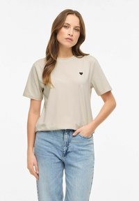 Beżowy bawełniany t-shirt z krótkimi rękawami, okrągłym dekoltem i małym czarnym logo w kształcie serca na lewej piersi. W zestawie z niebieskimi dżinsami z wysokim stanem.