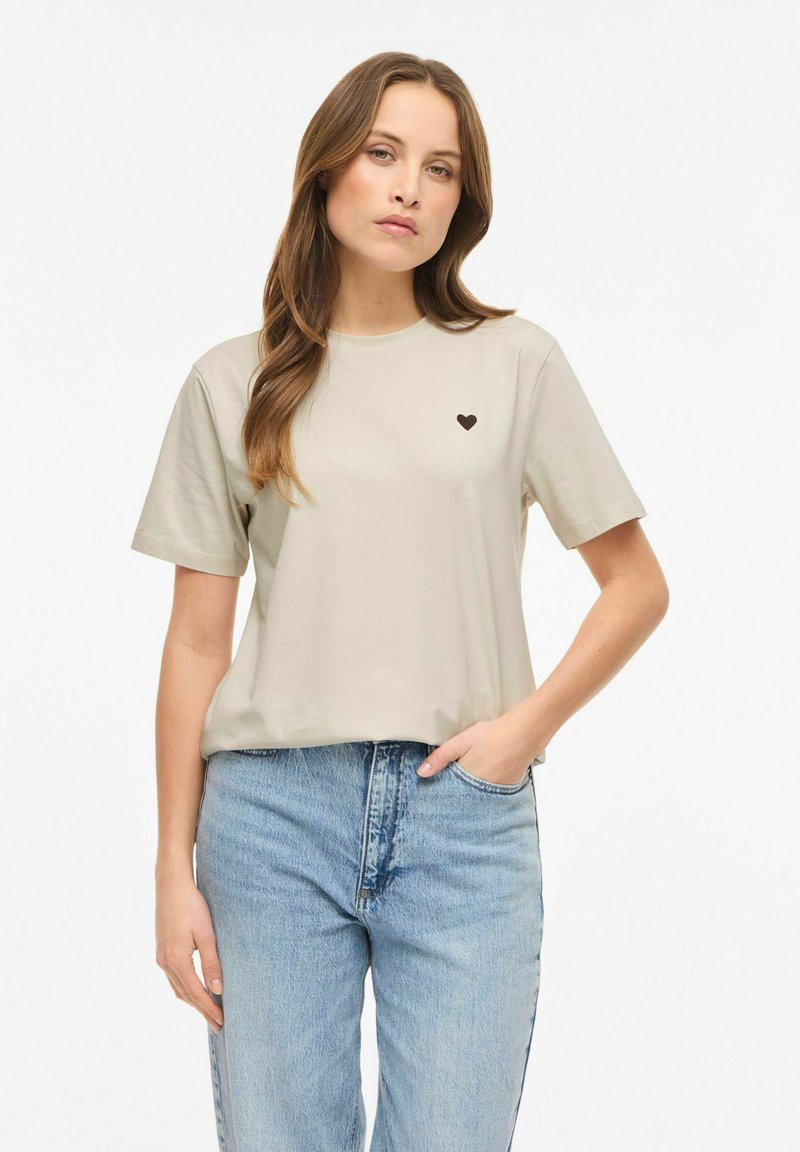 Beżowy bawełniany t-shirt z krótkimi rękawami, okrągłym dekoltem i małym czarnym logo w kształcie serca na lewej piersi. W zestawie z niebieskimi dżinsami z wysokim stanem.