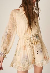 Mujer que lleva un vestido largo de manga larga con un ligero estampado floral, botones, cinturón en la cintura y volante en el dobladillo, posando frente a un fondo liso.