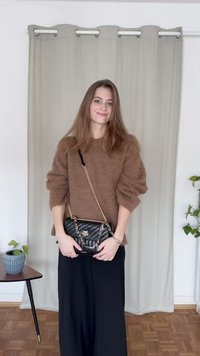 Brązowy, puszysty sweter, czarna pikowana torebka na łańcuszku, długie czarne spodnie, wnętrze z neutralną zasłoną i roślinami.