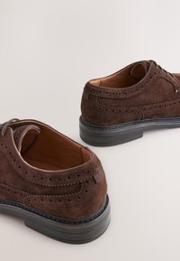 Chaussures décontractées en daim marron avec des détails brogue, bout rond et semelle en caoutchouc noire, présentant une texture lisse et une fermeture à lacets.