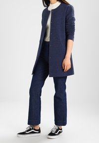 Marineblå lång cardigan med en texturerad design, framknäppningar och figursydda ärmar, parat med högmidjade mörka denimjeans och svarta sneakers.