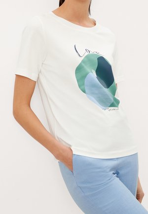 Frau trägt weißes Kurzarmshirt mit abstraktem grün-blauem Muster und Schriftzug "love", kombiniert mit hellblauen Hosen, Hand in der Tasche.