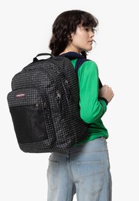 Mochila a cuadros en blanco y negro con bolsillo frontal de malla, doble tirador de cremallera, correas acolchadas y costuras reforzadas.