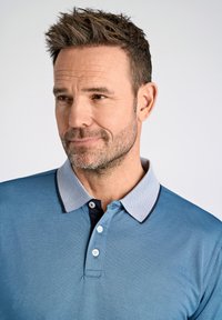 Blå polo med texturerad tyg, med en kontrasterande ljusblå mönstrad krage och treknapps öppenhet.