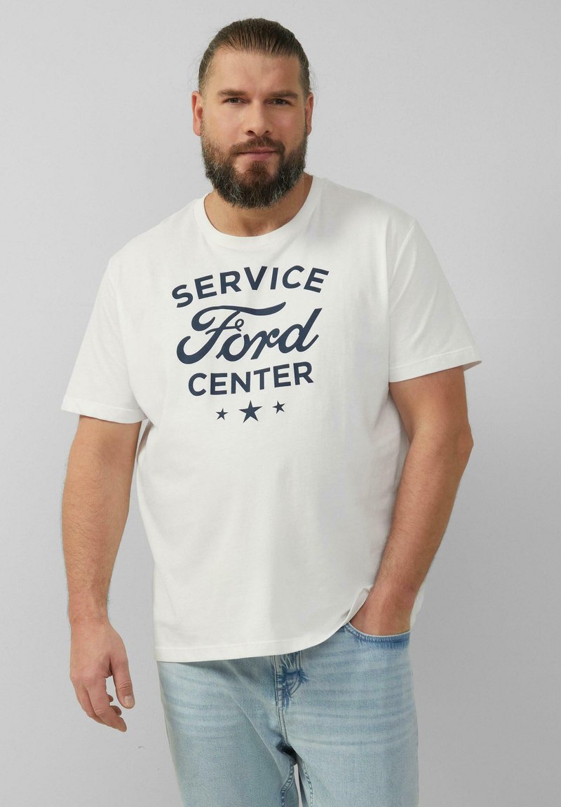 Baardman in een wit "Service Ford Center" T-shirt en lichtblauwe spijkerbroek, staande tegen een effen lichtgrijze achtergrond.