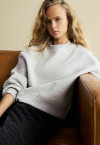 Grauer Pullover mit weicher Textur, übergroßer Passform und gerippten Manschetten, kombiniert mit dunklen strukturierten Hosen, sitzend auf einem braunen Ledersofa.