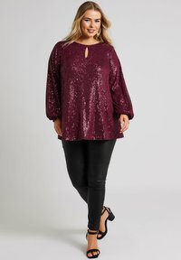 Burgundy paljettopp med rund hals och nyckelhålsdetalj, kombinerad med svarta leggings och svarta remsandaler; långa, puffiga ärmar.