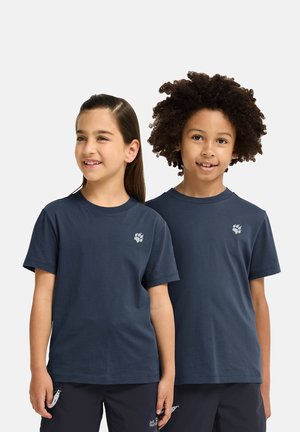 Deux enfants souriants portant des t-shirts bleu marine assortis avec des logos d'empreintes de pattes et un pantalon noir, debout côte à côte sur un fond clair.