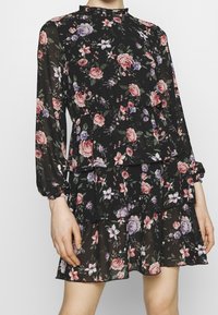 Robe fleurie en tissu noir ornée de roses roses et violettes, manches longues, encolure à volants et jupe à étages.