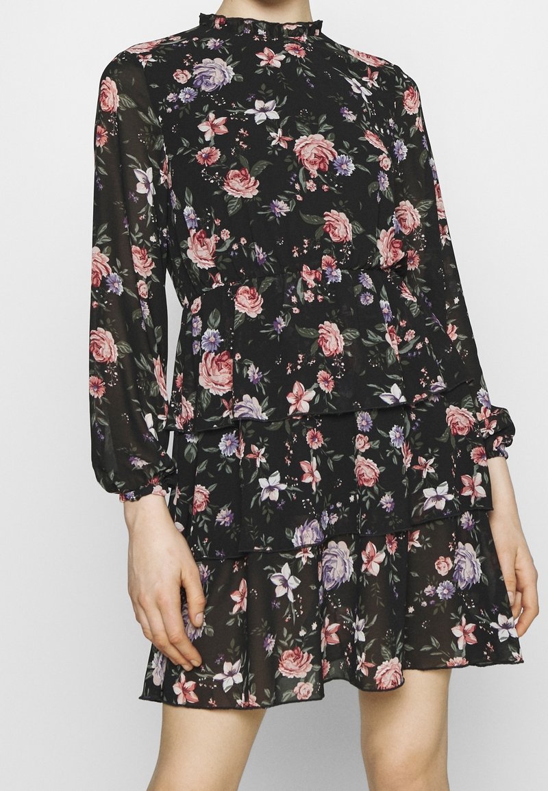 Robe fleurie en tissu noir ornée de roses roses et violettes, manches longues, encolure à volants et jupe à étages.