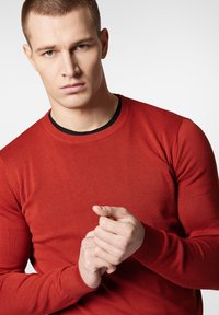 Czerwony sweter dziany z ściągaczem przy dekolcie, dopasowany fason i teksturowany materiał. Ręce modela są złożone przed nim.