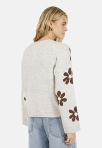 Pull en maille gris clair avec des motifs floraux bruns sur les manches, des poignets côtelés et une coupe décontractée. La surface texturée ajoute de la profondeur.