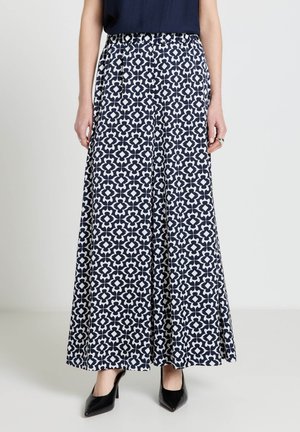 Donna che indossa pantaloni a gamba larga con motivo geometrico blu navy e bianco, abbinati a tacchi neri a punta e una canottiera scura senza maniche.