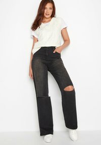 Sorte slidte wide-leg jeans med hulkeskader på knæene, parret med en hvid t-shirt og en cremefarvet kabelstrikket vest. Stylet med hvide sneakers.