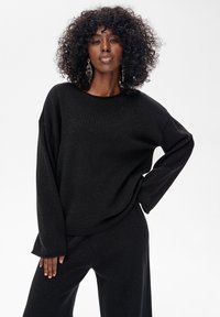 Czarny dziergany sweter o luźnym kroju, z długimi rękawami i zaokrąglonym dekoltem. Tkanina ma subtelny połysk, zestawiona z pasującymi czarnymi spodniami.