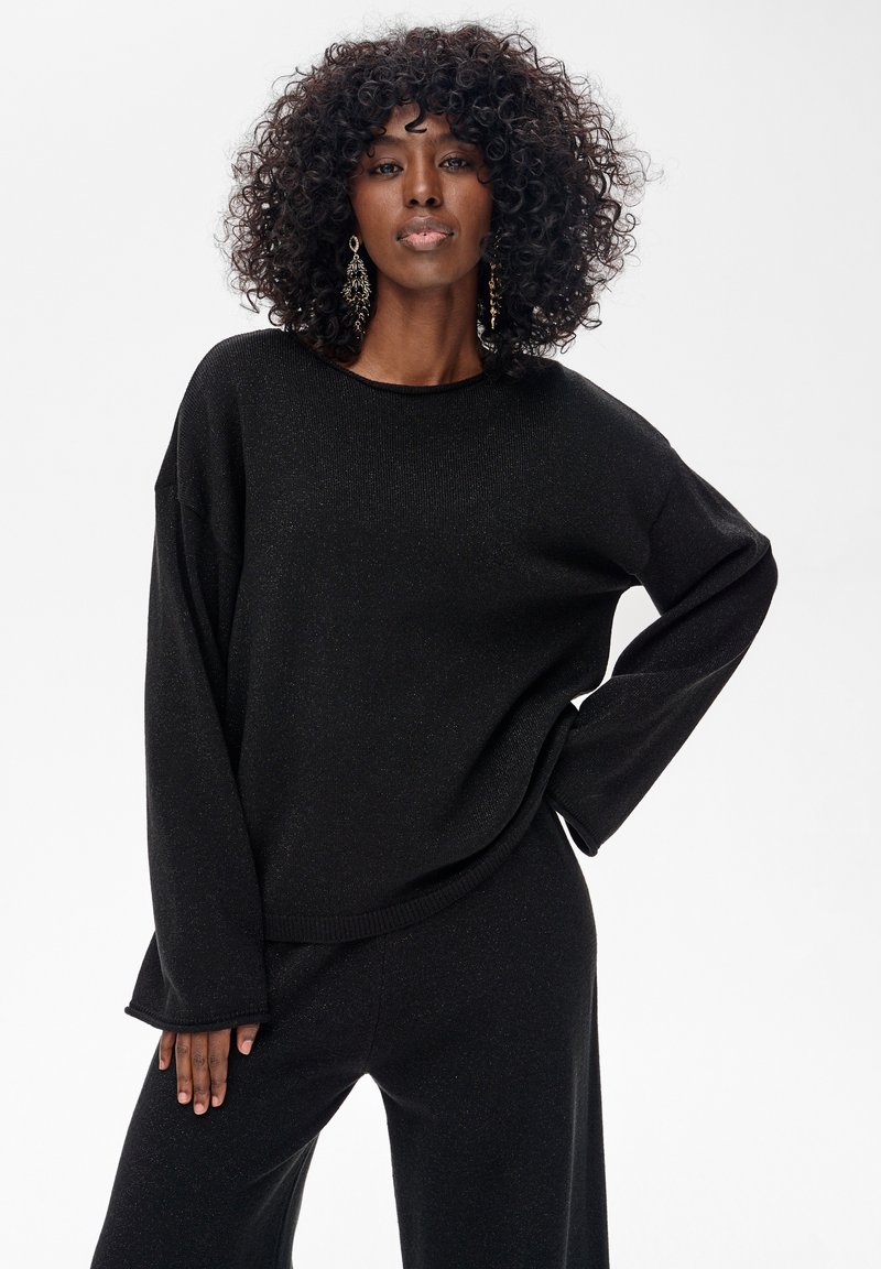 Czarny dziergany sweter o luźnym kroju, z długimi rękawami i zaokrąglonym dekoltem. Tkanina ma subtelny połysk, zestawiona z pasującymi czarnymi spodniami.