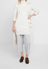 Kvinna som bär en krämfärgad poloklänning, ljusa rutiga korta byxor, en krämfärgad trenchcoat och bruna loafers, stående mot en vit bakgrund.