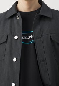 Svart skjortjacka med ränder och silverknappar samt fickor, lager över en svart t-shirt med en turkos grafisk design.